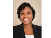 Smiling woman in black blazer, studio headshot on beige background.