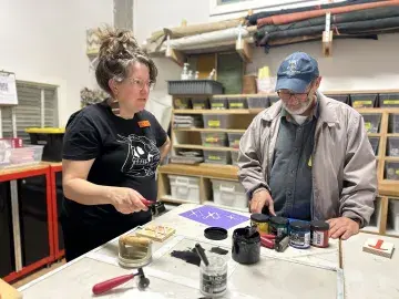 Two people at a workshop bench assembling parts with tools and a coffee mug.