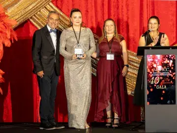 Five people on stage with red curtain with Karen in silver dress holds award by podium.