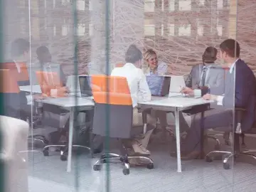 Business meeting in a glass-walled office, people at a table with laptops and orange chairs