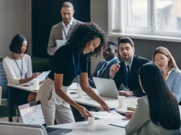 Team meeting in office; woman leans over table reviewing documents with colleagues
