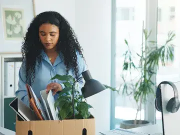 Woman with curly hair packing office items into a cardboard box in a bright workspace.