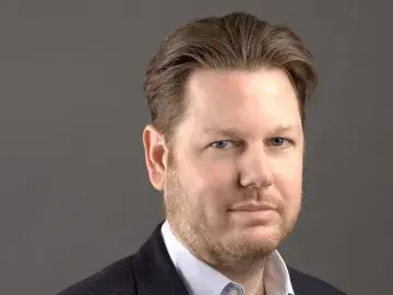 Studio portrait of a man in a navy blazer and white shirt against a gray background.