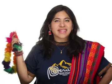 Smiling woman holding a colorful feathered item, wearing a woven sash on white background.