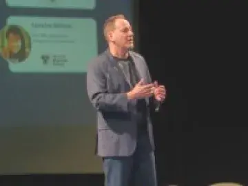 Man in blazer speaking on stage in front of projected slide and large “TEDx” letters