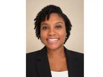 Smiling woman in black blazer, studio headshot on beige background.