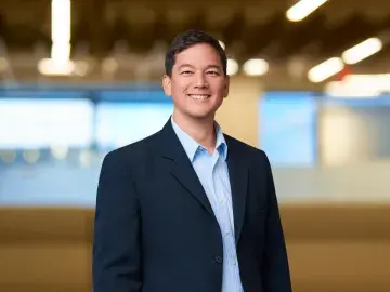 Smiling man in navy blazer and blue shirt standing in a parking garage, hands in pockets.
