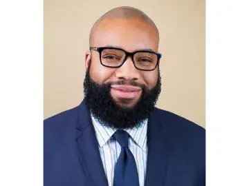 Eben Smith with glasses and full beard in a navy suit against a beige background