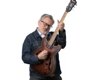 Graham Dodge with glasses holding an electric guitar on a white background.