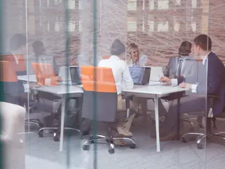 Business meeting in a glass-walled office, people at a table with laptops and orange chairs