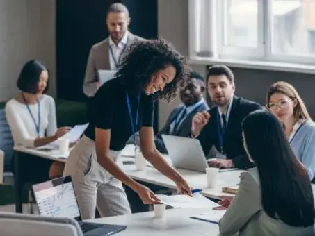 Team meeting in office; woman leans over table reviewing documents with colleagues