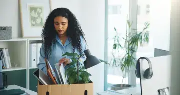 Woman with curly hair packing office items into a cardboard box in a bright workspace.
