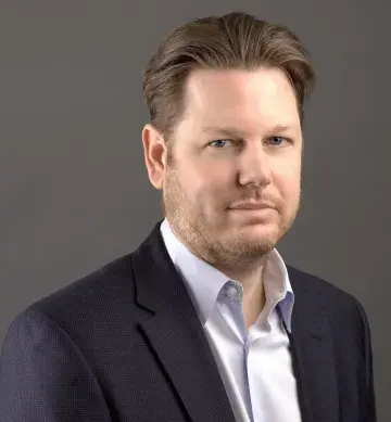 Studio portrait of a man in a navy blazer and white shirt against a gray background.