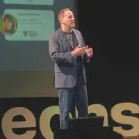 Man in blazer speaking on stage in front of projected slide and large “TEDx” letters