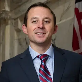 Portrait photo of a smiling man in a suit and striped tie, flag in background.
