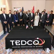 Group in suits posing behind TEDCO table banner in conference room with flags.